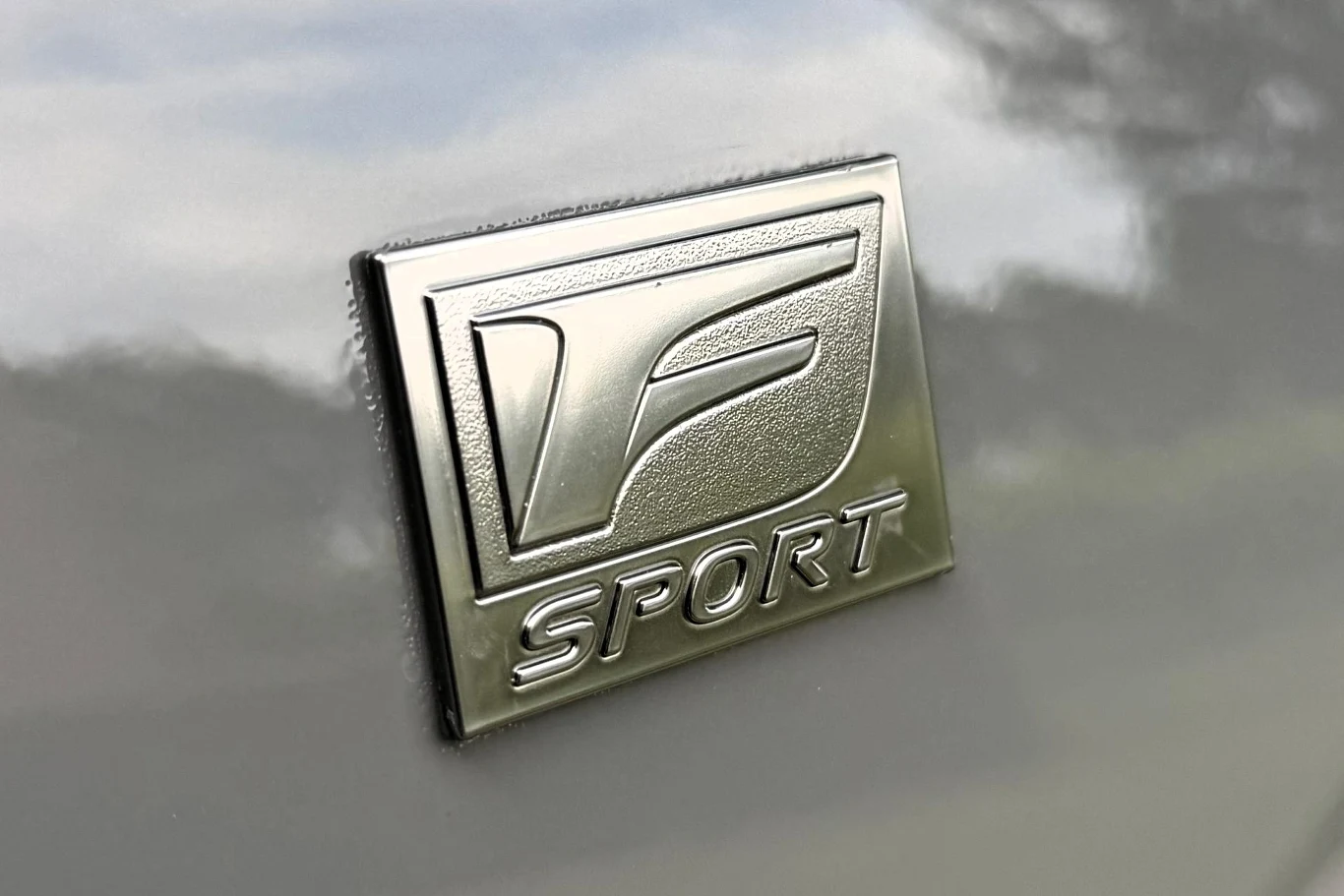Lexus NX w wariancie F SPORT wyróżnia się m.in. 20-calowymi felgami aluminiowymi ze specjalnym wzorem i czerwonymi zaciskami. Chromowany emblemat z literą F i napisem SPORT umieszczony na jasnej, metalicznej powierzchni.