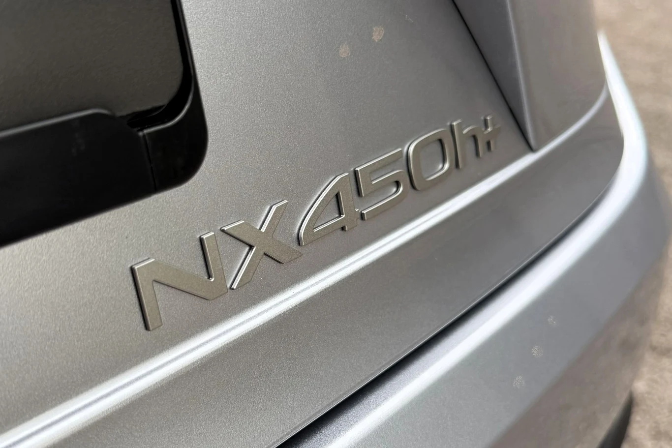 Lexus NX z hybrydą plug-in ma przejeżdżać ponad 100 km na jednym ładowaniu. Srebrny znaczek NX450h+ umieszczony na tylnej klapie samochodu w kolorze szarym.