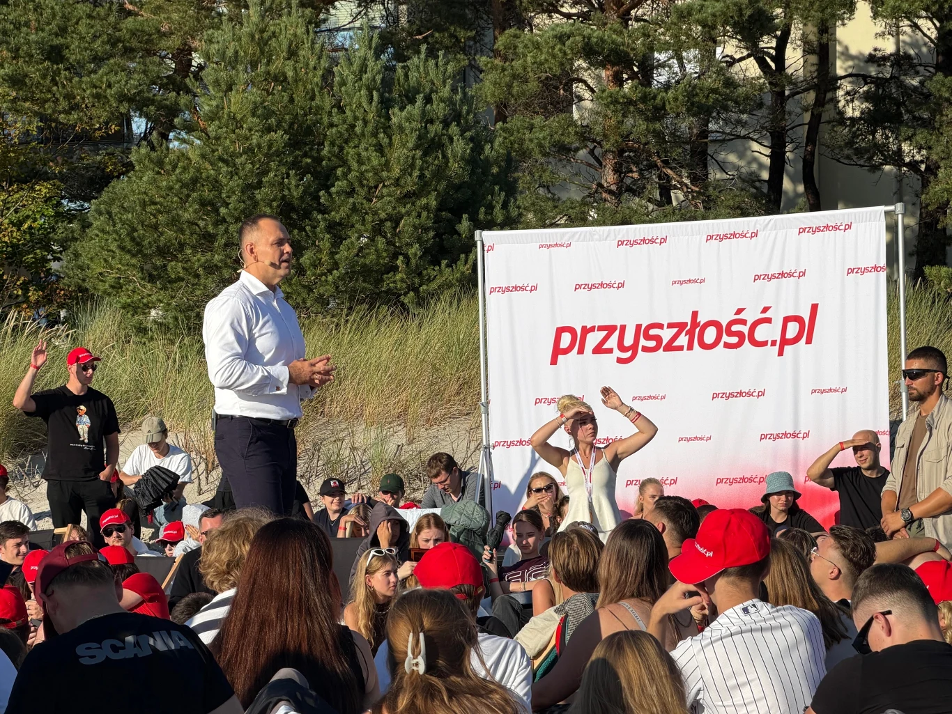 Karol Nawrocki podczas sesji pytań i odpowiedzi Mężczyzna w białej koszuli przemawia do licznej grupy ludzi zgromadzonej na tle baneru z napisem „przyszłość.pl”, w otoczeniu zielonych drzew i trawiastych wydm.