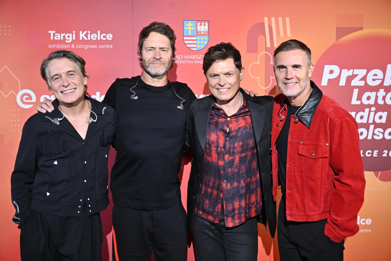 Take That i Michael Patrick Kelly (drugi z prawej) podczas Magicznego Zakończenia Wakacji w Kielcach Czterech mężczyzn pozuje wspólnie na tle czerwonej ścianki z logotypami wydarzenia, ubrani w stylizacje sceniczne; dwóch z nich ma na sobie czarne ubrania, jeden czerwoną kurtkę, a jeden czarną marynarkę z czerwono-czarną koszulą.