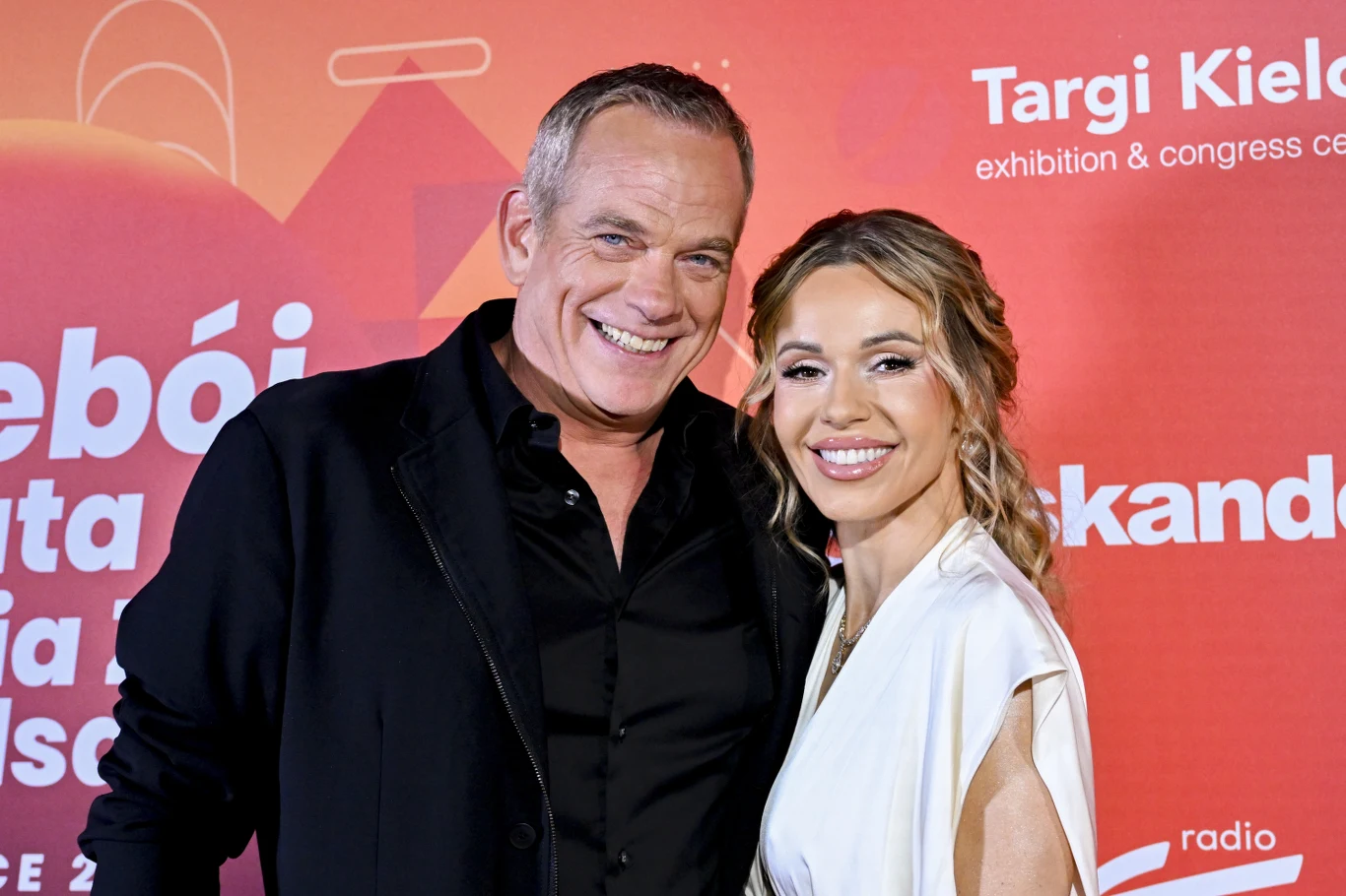 Garou i Doda wystąpili razem w duecie w Kielcach Uśmiechnięty mężczyzna w czarnej marynarce i kobieta w białej sukni stoją razem na tle ścianki z czerwonymi napisami wydarzenia.