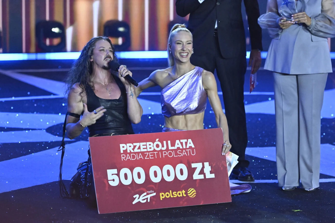 Triumfujący Michał Szpak z prowadzącą Idą Nowakowską Michał Szpak i Ida Nowakowska na scenie z tablicą 'Przebój Lata 500 000 zł', wśród elegancko ubranych osób.