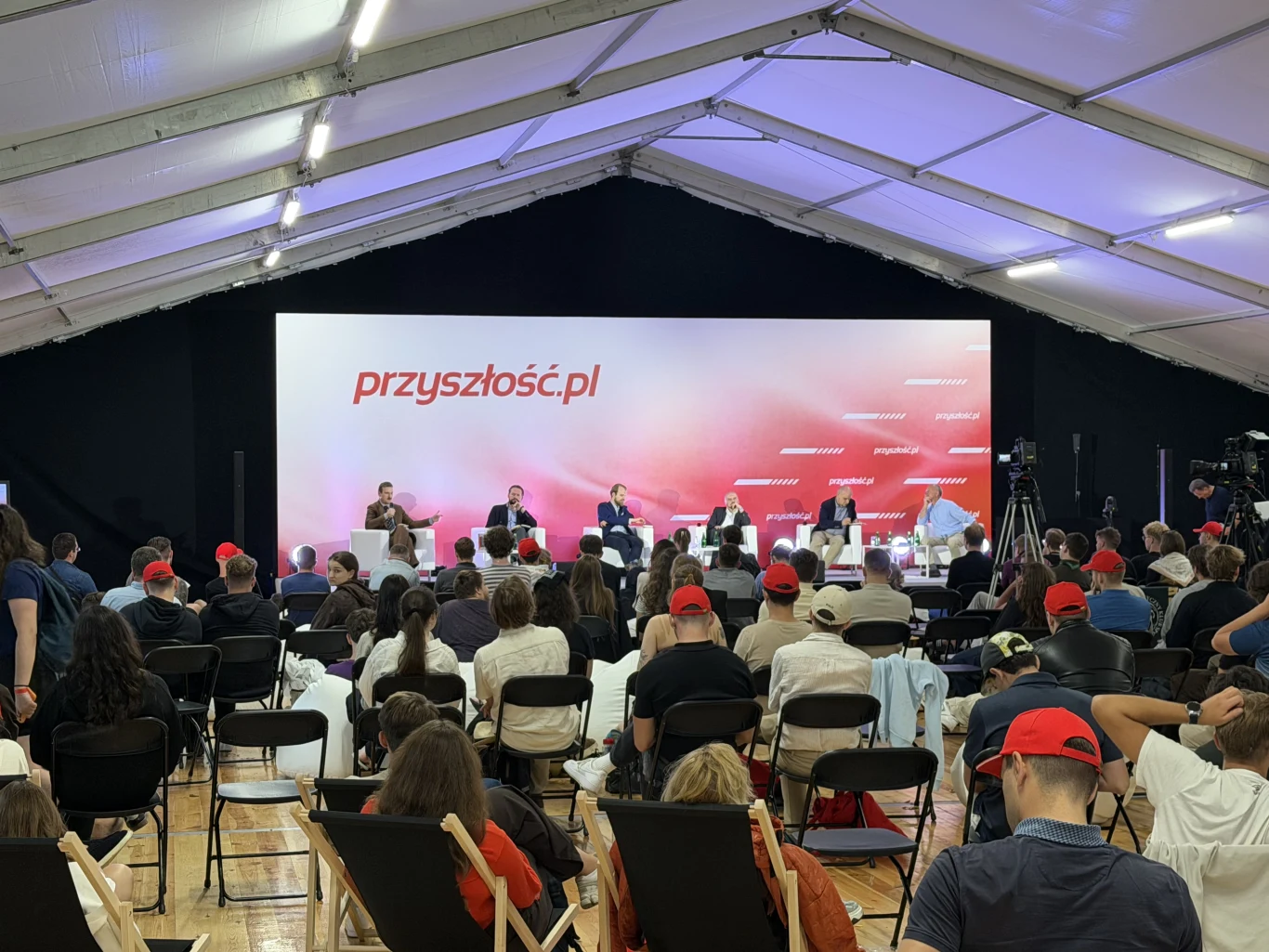 Strefa paneli tematycznych podczas wydarzenia "Przyszłość.pl" Panel dyskusyjny z udziałem sześciu osób na scenie, moderowanych przez prowadzącego, skierowanych do licznej publiczności siedzącej na składanych krzesłach i leżakach pod białym namiotem, na tle dużego baneru z napisem 'przyszłość.pl'.
