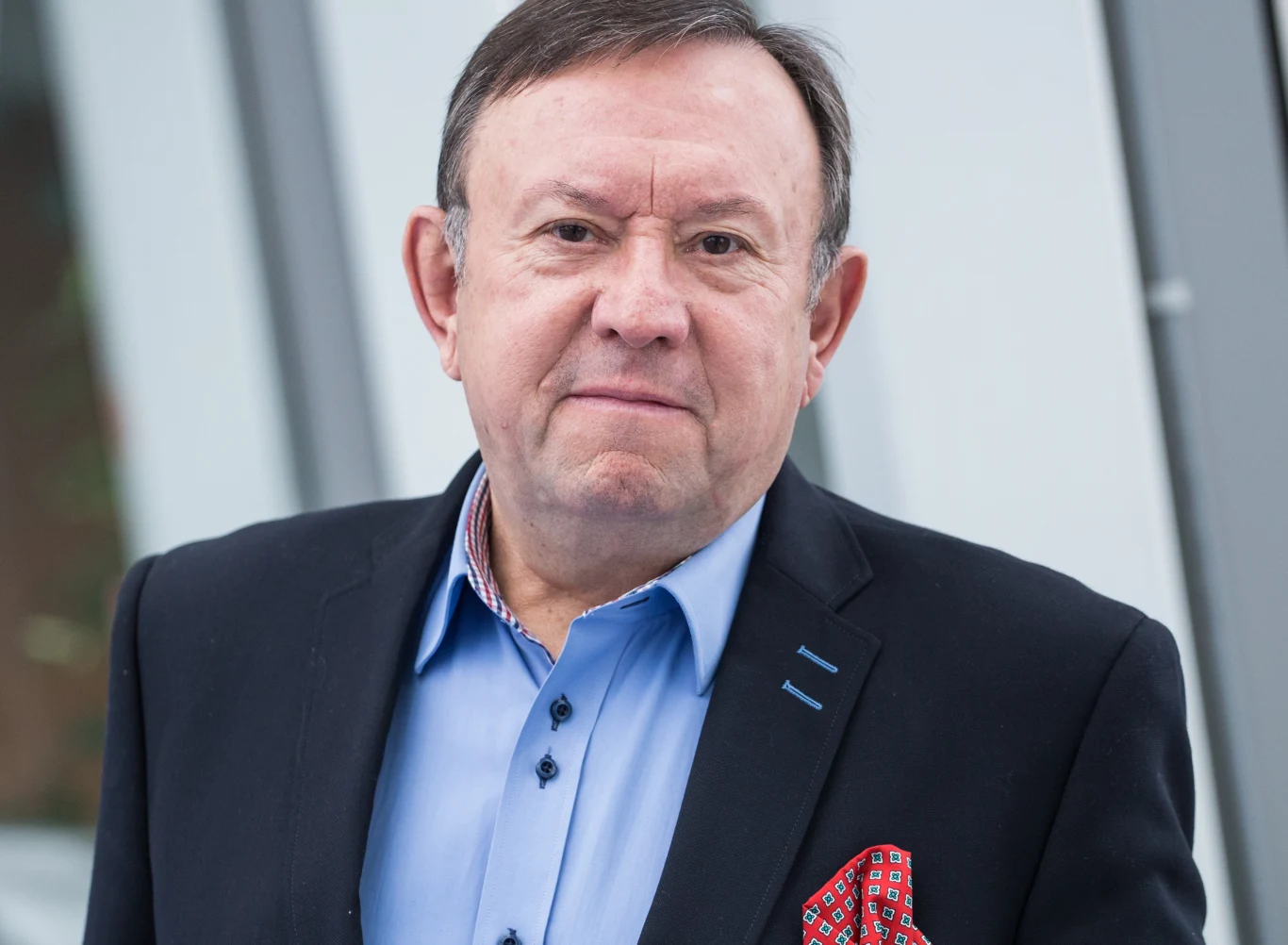 Zbigniew Buczkowski Zbigniew Buczkowski w marynarce, błękitnej koszuli i poszetce, stoi przy przeszklonej ścianie.