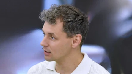 Mężczyzna o krótkich, lekko kręconych włosach i jasnej karnacji ubrany w białą, prążkowaną koszulkę polo z trzema guzikami, zwrócony lekko bokiem, na rozmytym tle.