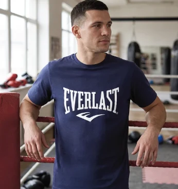 Mężczyzna w granatowej koszulce z napisem Everlast opiera się o linę ringu bokserskiego w dobrze oświetlonej sali treningowej.