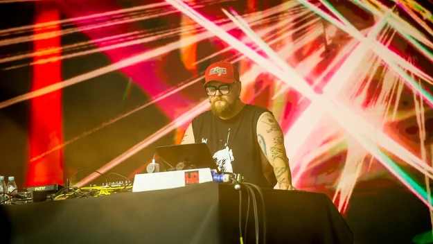 Brodaty mężczyzna w czapce i okularach obsługuje sprzęt DJ-ski na tle kolorowych, dynamicznych świateł i laserów.