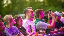 Grupa ludzi przy scenie na koncercie, skupionych na występie, trzymających telefony komórkowe i robiących zdjęcia, oświetlona kolorowym światłem.