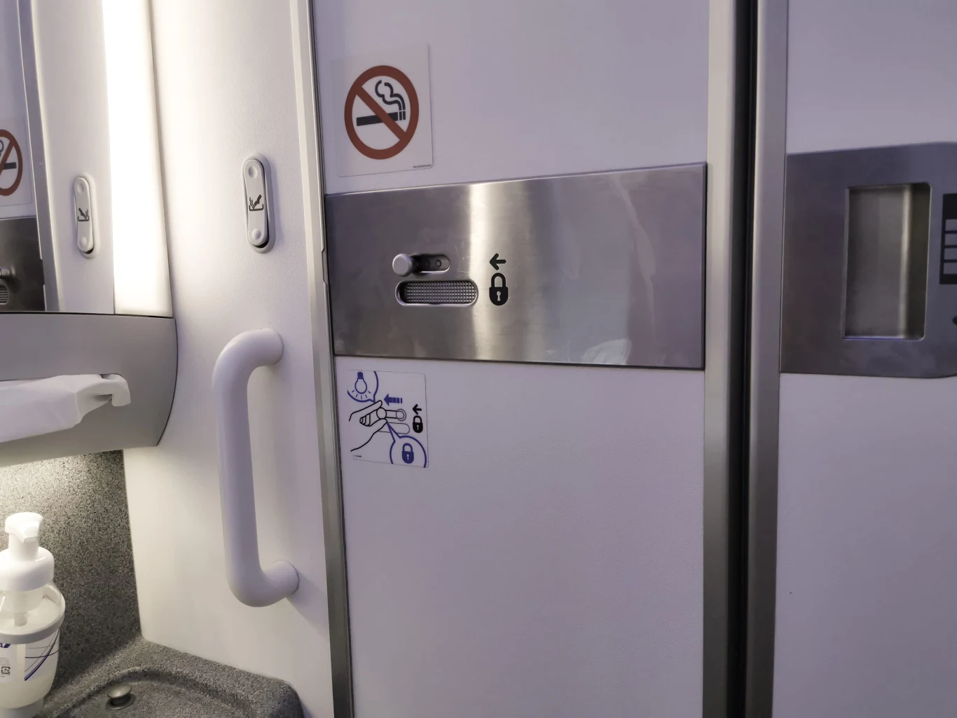 Drzwi toalety samolotu z widocznym zamkiem, uchwytem, dozownikiem płynu oraz symbolem zakazu palenia.