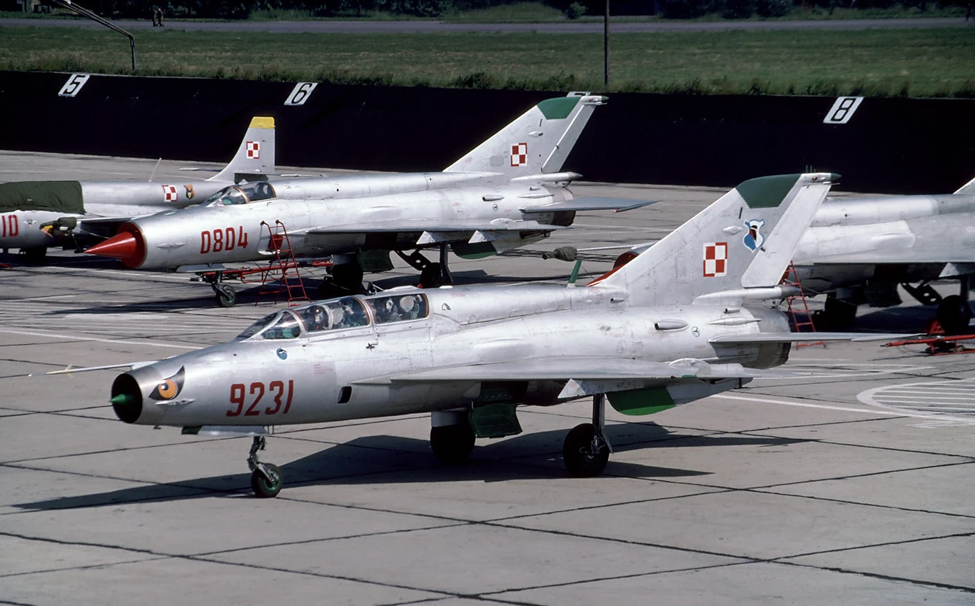 Dwa polskie myśliwce odrzutowe Su-7 o metalicznym kolorze stojące na lotniskowej płycie postojowej, każdy z oznaczeniami państwowymi na statecznikach pionowych.