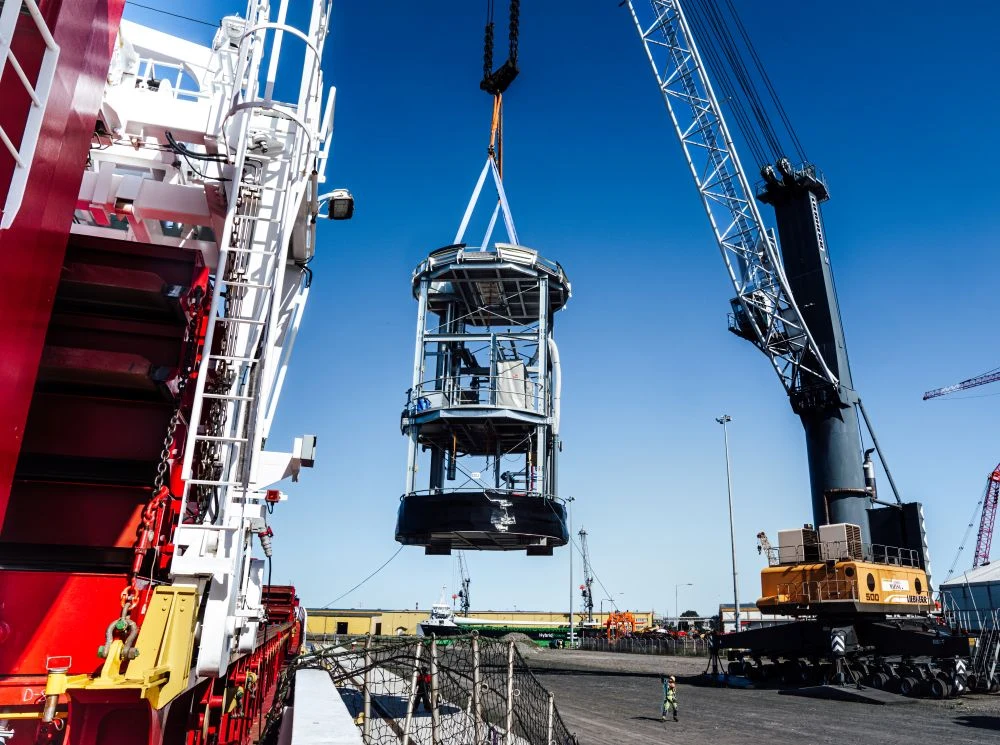 Smulders Projects Poland Duży dźwig transportuje metalową konstrukcję nad pokładem statku w porcie, w tle widoczne są maszyny i sprzęt przeładunkowy.
