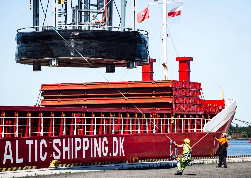 Smulders Projects Poland Czerwony statek towarowy BALTIC SHIPPING z załadunkiem unoszonym dźwigiem, dwóch pracowników portowych obsługujących liny przy nabrzeżu.