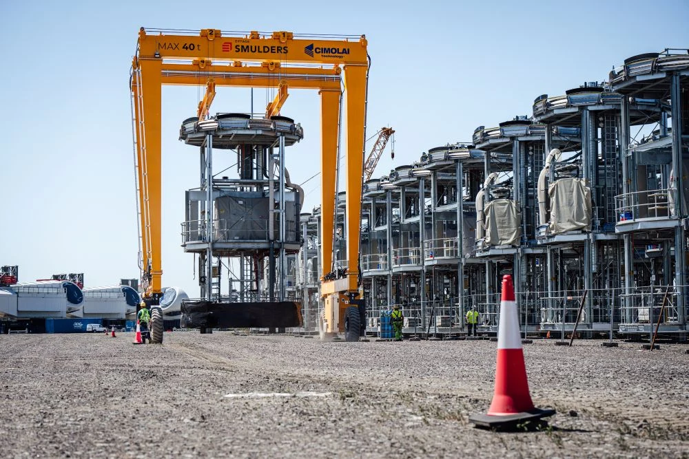 Smulders Projects Poland Ogromny żółty dźwig transportuje metalową konstrukcję, podczas gdy po prawej stronie ustawione są rzędy podobnych stalowych elementów na placu przemysłowym.