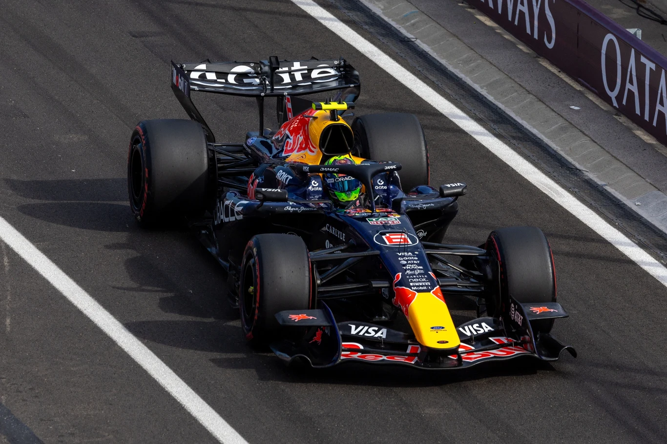 Isack Hadjar Samochód wyścigowy Red Bull Racing poruszający się po torze, kierowca w żółtym kasku w kokpicie, widoczne logo sponsora na tylnej osi i elementach karoserii.