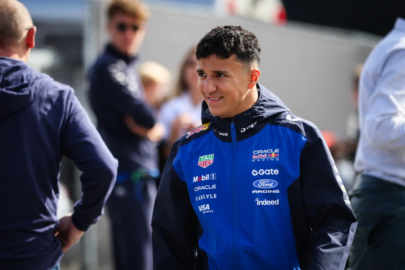 Isack Hadjar Młody mężczyzna w niebieskiej kurtce zespołu Oracle Red Bull Racing stoi na świeżym powietrzu i rozmawia z inną osobą, w tle kilka nieostrych postaci.