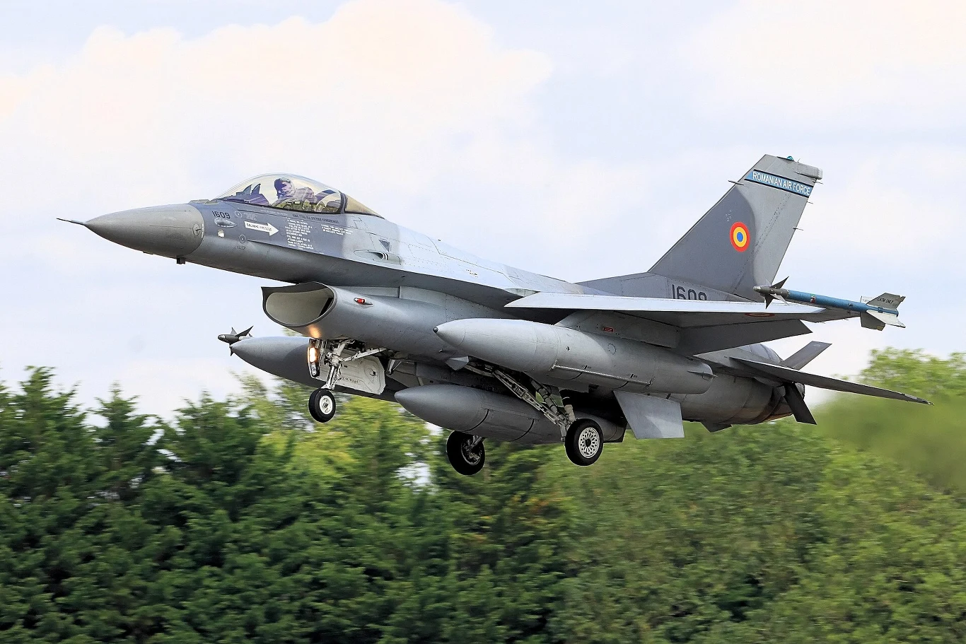 Myśliwiec F-16 w barwach rumuńskich sił powietrznych w trakcie podchodzenia do lądowania na tle zielonych drzew i nieba.