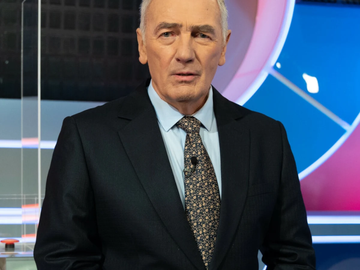 Karol Strasburger Karol Strasburger w garniturze z krawatem na tle scenografii telewizyjnej programu Familiada.
