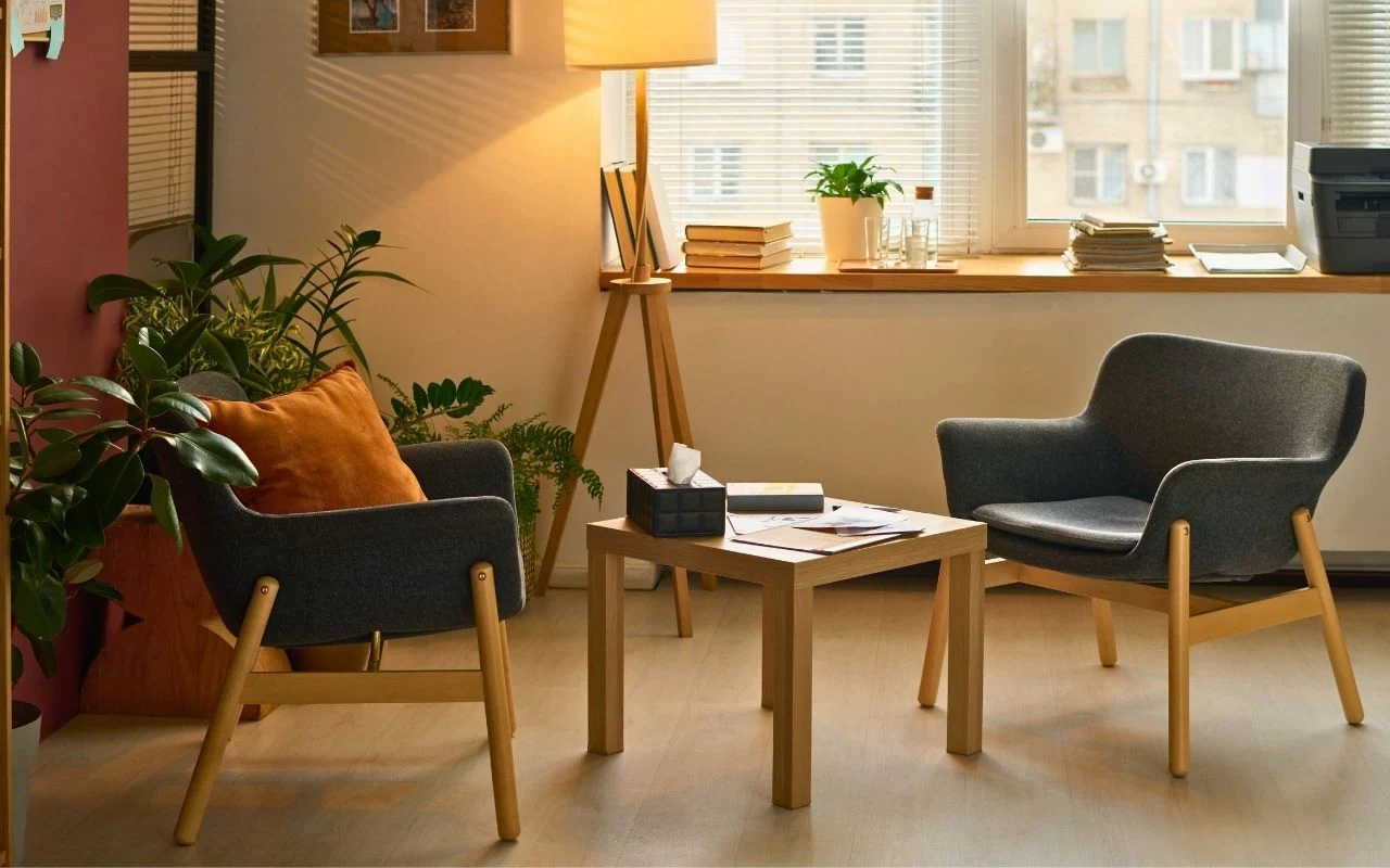 Dwa designerskie fotele z drewnianymi nogami ustawione wokół małego stolika z chusteczkami i książkami, w jasnym, przytulnym wnętrzu z dużymi oknami i zielonymi roślinami.