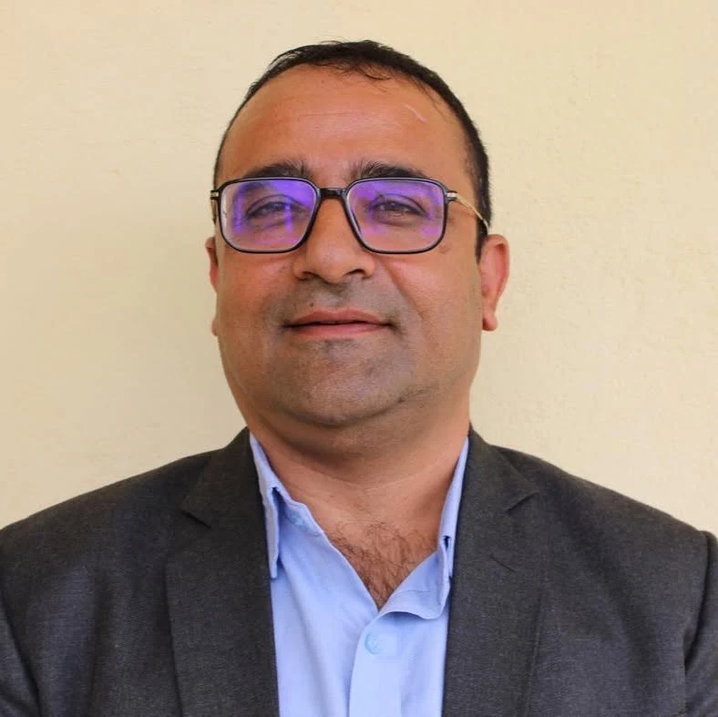 Rajendra Dawadi, dyrektor szkoły średniej Tribhuvan Trishuli, ewakuowanej w ostatniej chwili przed nadejściem fali powodziowej Mężczyzna w okularach i jasnoniebieskiej koszuli pod ciemną marynarką, patrzący przed siebie na tle jasnej, jednolitej ściany.