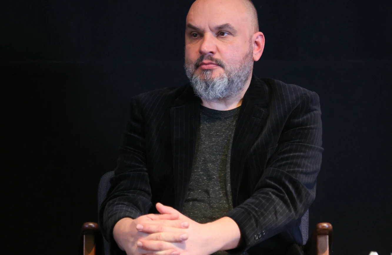 Witold Płóciennik na przeglądzie Polskie Nagrody Filmowe Orły 2022 Witold Płóciennik siedzi na krześle, w ciemnej marynarce i szarej koszulce, na tle czarnej ściany.