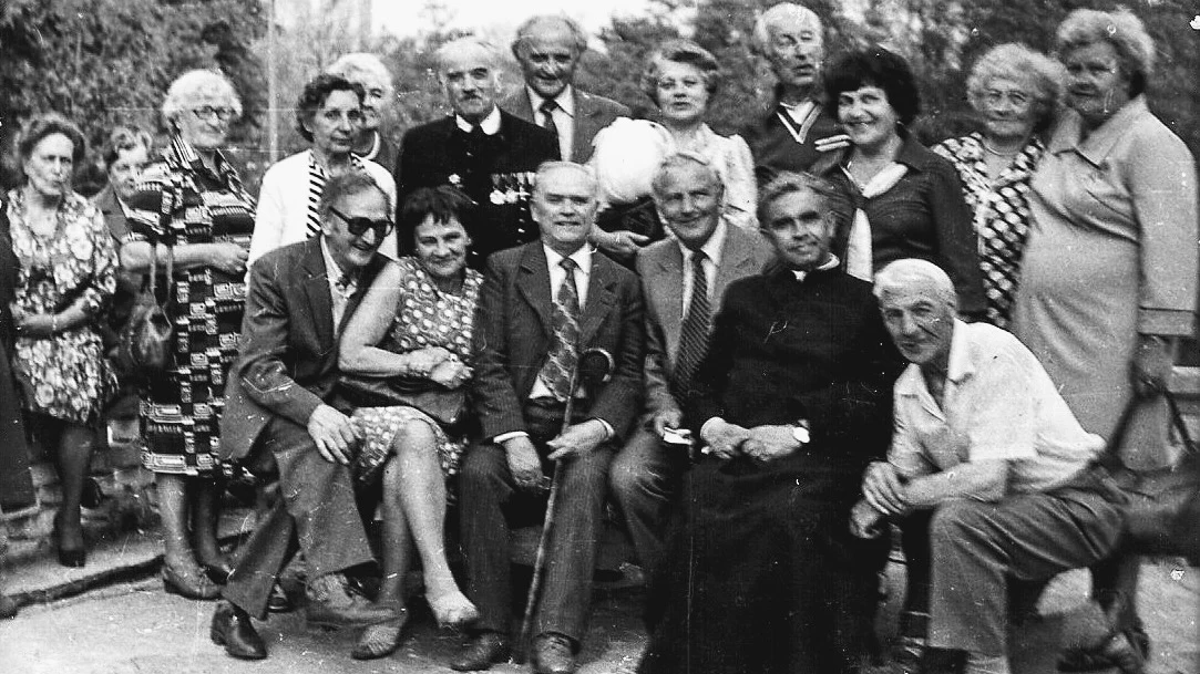 Grupa kilkunastu starszych osób, w tym mężczyźni i kobiety ubrani odświętnie, zgromadzona na zewnątrz w ogrodzie lub parku, część z nich siedzi, a pozostali stoją, tworząc zwartą, zżywioną grupę.