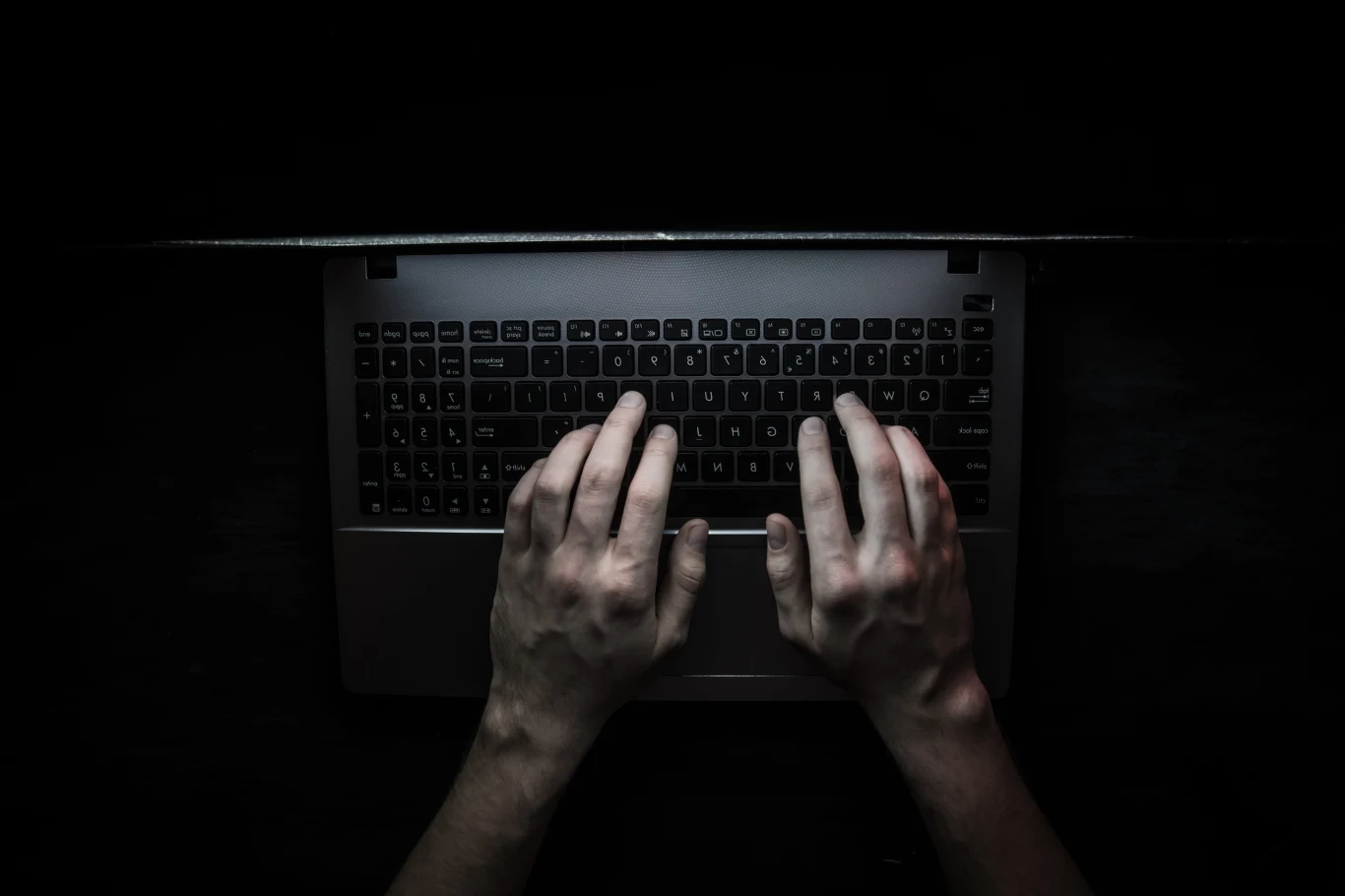 Zdjęcie ilustracyjne Dwie dłonie na podświetlonej klawiaturze laptopa w ciemnym otoczeniu.
