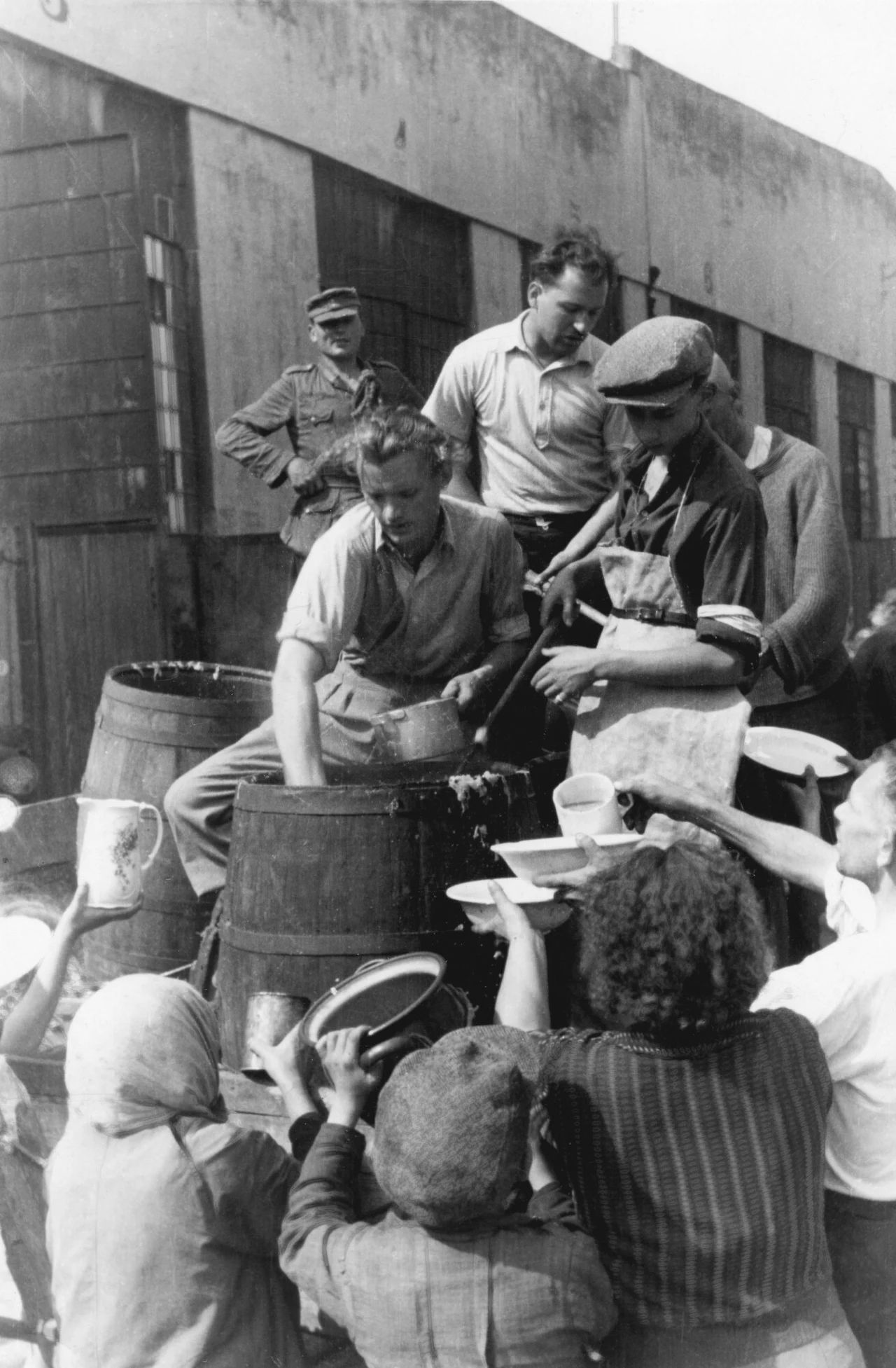 Rozdawanie jedzenia w trakcie Powstania Warszawskiego, wrzesień 1944 Grupa ludzi ustawia się przy beczkach, do których nalewany jest prawdopodobnie posiłek, podczas gdy kilku mężczyzn rozdziela żywność do misek i kubków wyciąganych przez tłum.