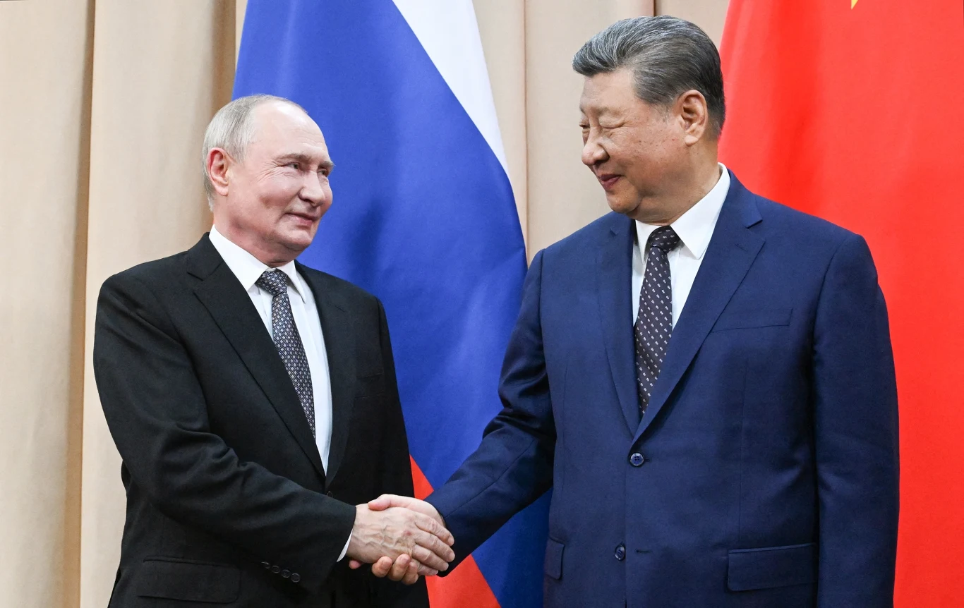 Władimir Putin spotkał się z Xi Jinpingiem Władimir Putin i Xi Jinping podają sobie ręce na tle flag Rosji i Chin, ubrani w garnitury.