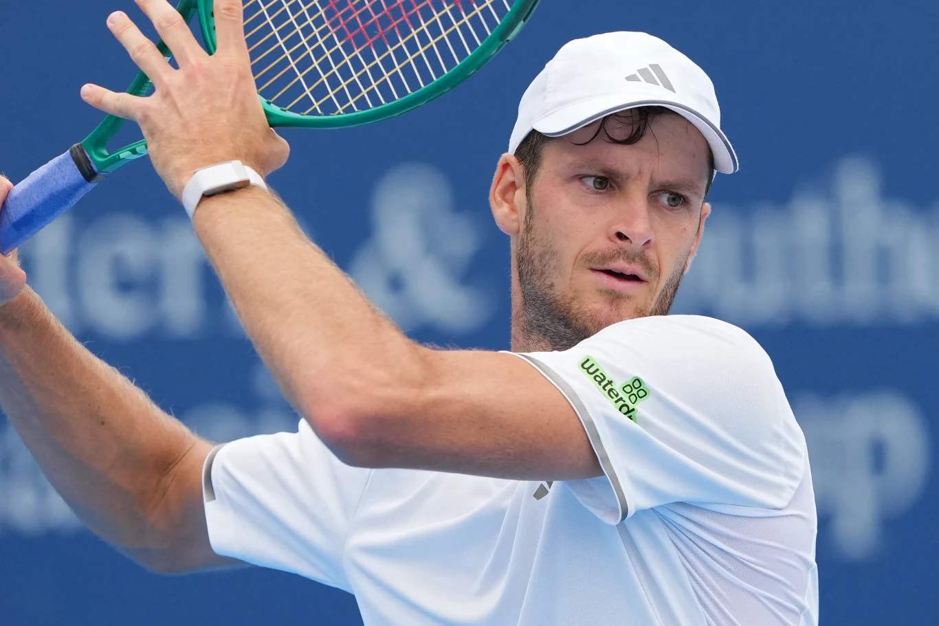 Hubert Hurkacz rywalizował z Damirem Dzumhurem o awans do drugiej rundy US Open Mężczyzna w białej koszulce i czapce trzyma rakietę tenisową, przygotowując się do uderzenia piłki.
