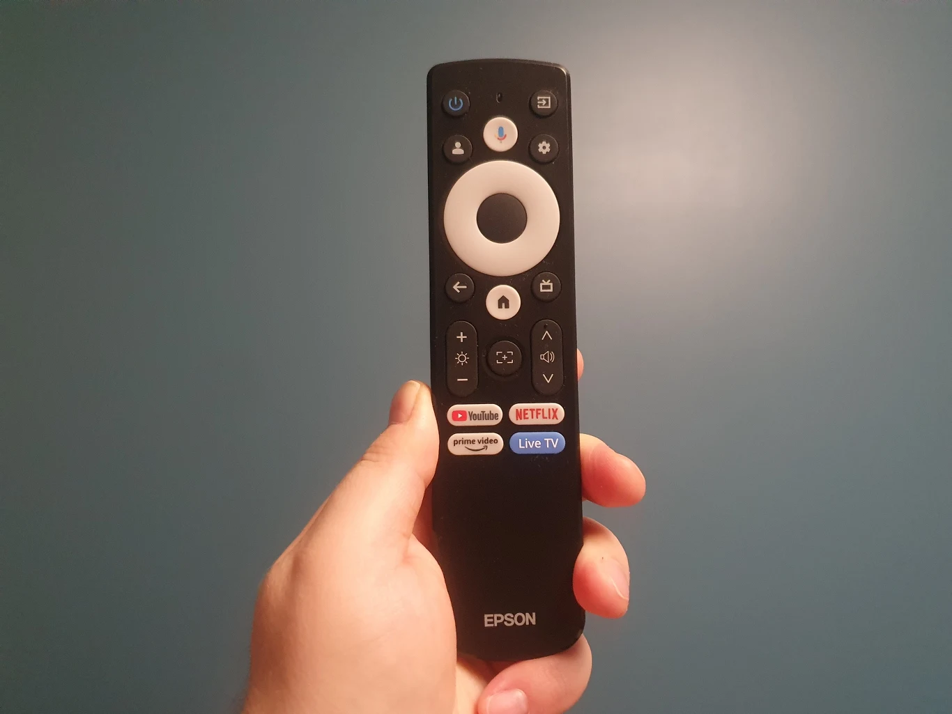 Pilot marki Epson z dużym białym przyciskiem centralnym oraz dedykowanymi przyciskami do aplikacji YouTube, Netflix i Live TV, trzymany w lewej ręce na tle jednolitej, niebieskiej ściany.