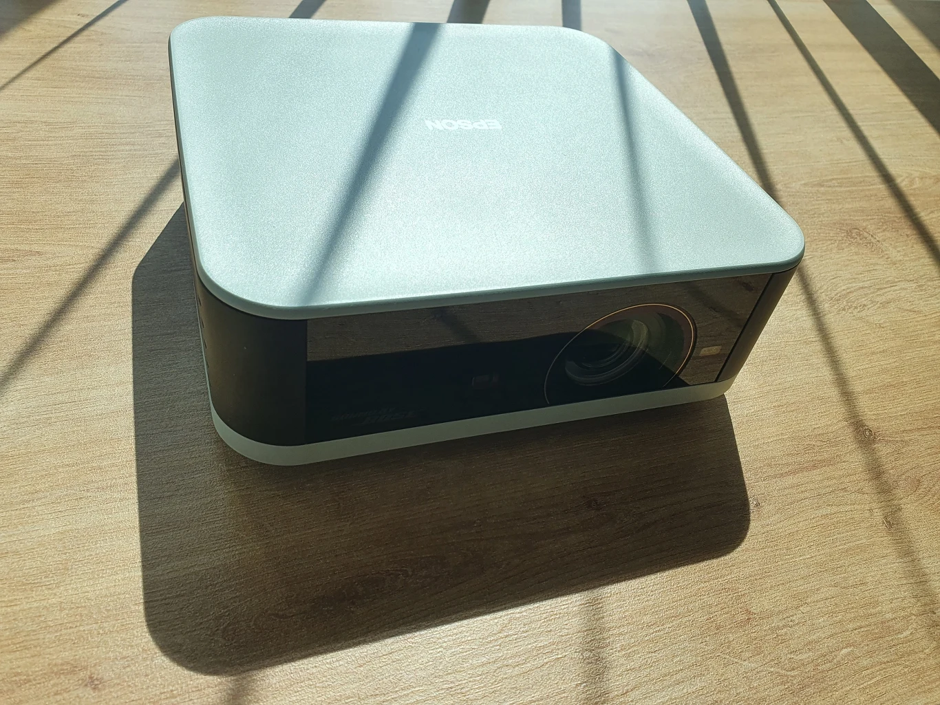 Nowoczesny projektor o minimalistycznym designie ustawiony na drewnianym blacie, oświetlany promieniami słońca, z wyraźnie widocznym obiektywem z przodu.