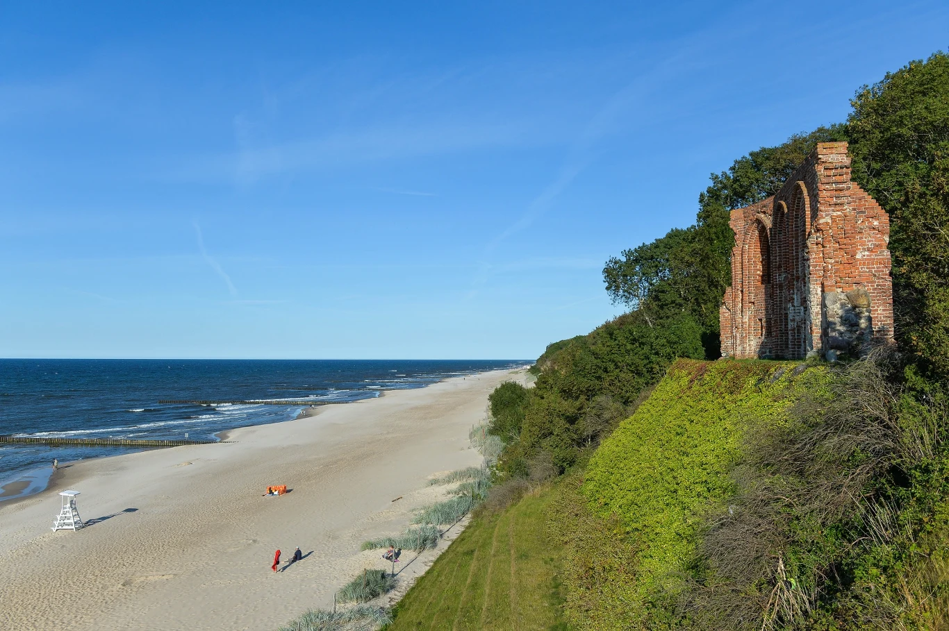Ruiny ceglanej budowli górujące nad piaszczystą plażą rozciągającą się wzdłuż brzegu morza, otoczone zielenią i widoczne na tle błękitnego nieba.