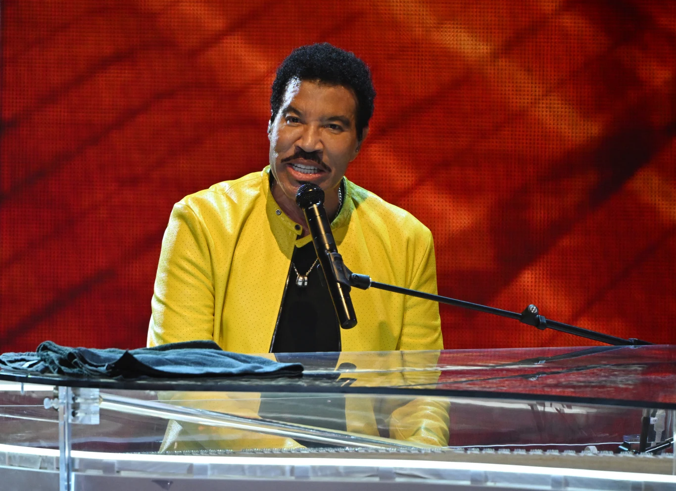 Lionel Richie Lionel Richie podczas koncertu: mężczyzna w żółtej kurtce gra na przezroczystym fortepianie i śpiewa do mikrofonu.