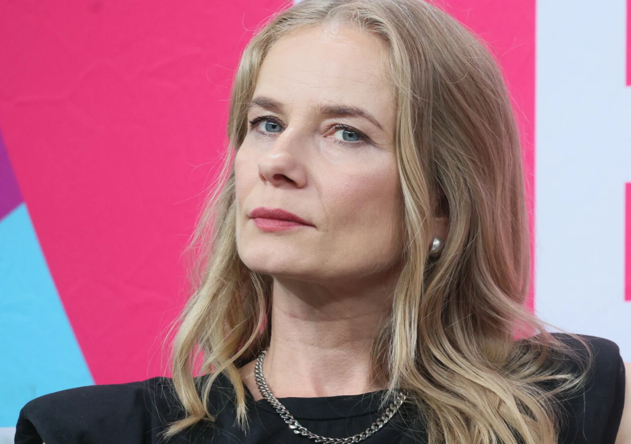 Magdalena Cielecka Kobieta o blond włosach, ubrana w czarną marynarkę i srebrny łańcuszek, patrzy poważnie przed siebie na tle kolorowej ściany.