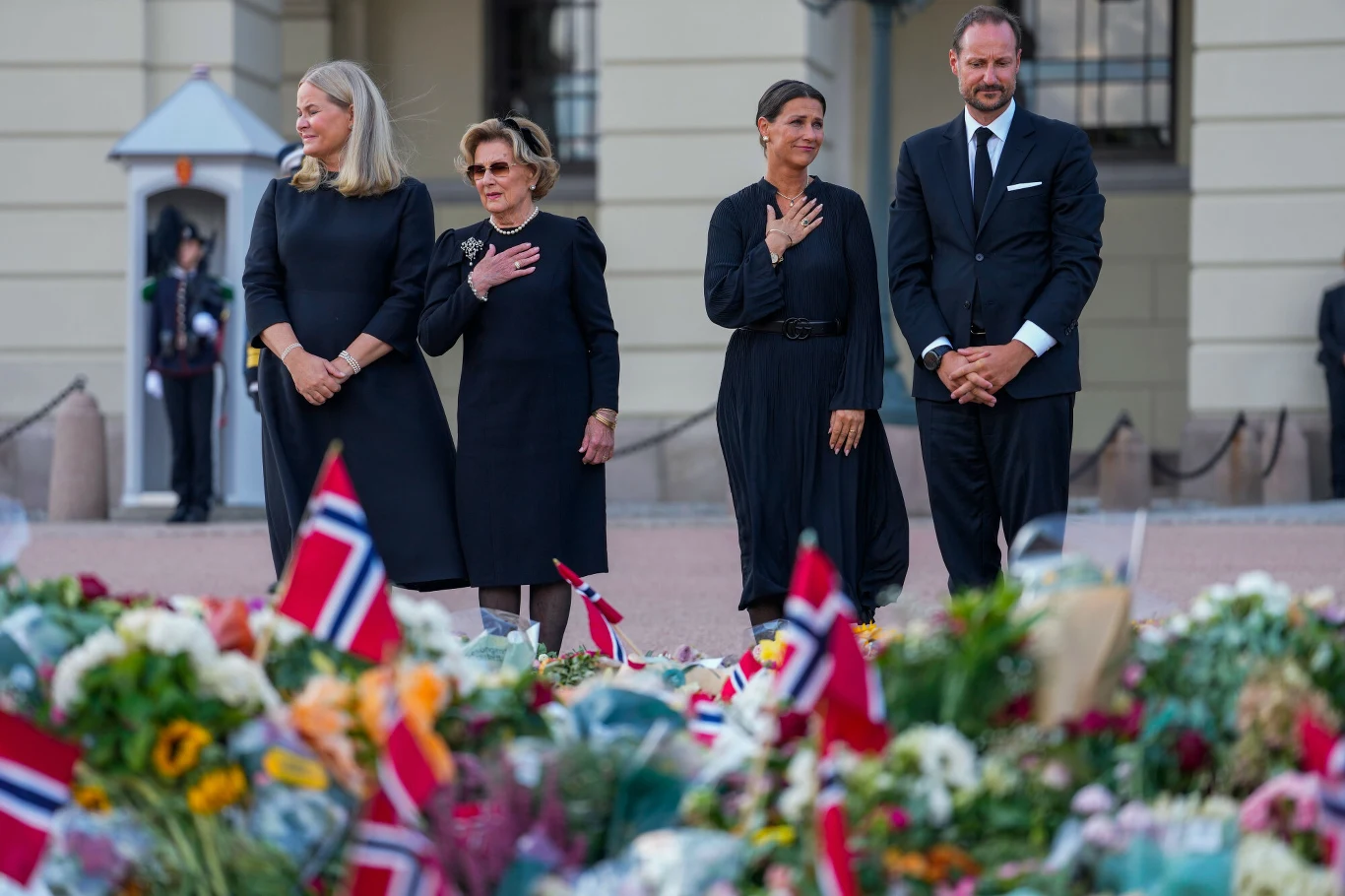 Cztery osoby ubrane w ciemne stroje stoją w skupieniu przed dużą liczbą kwiatów i norweskich flag ułożonych na ziemi, wyrażając szacunek i powagę w miejscu upamiętnienia.