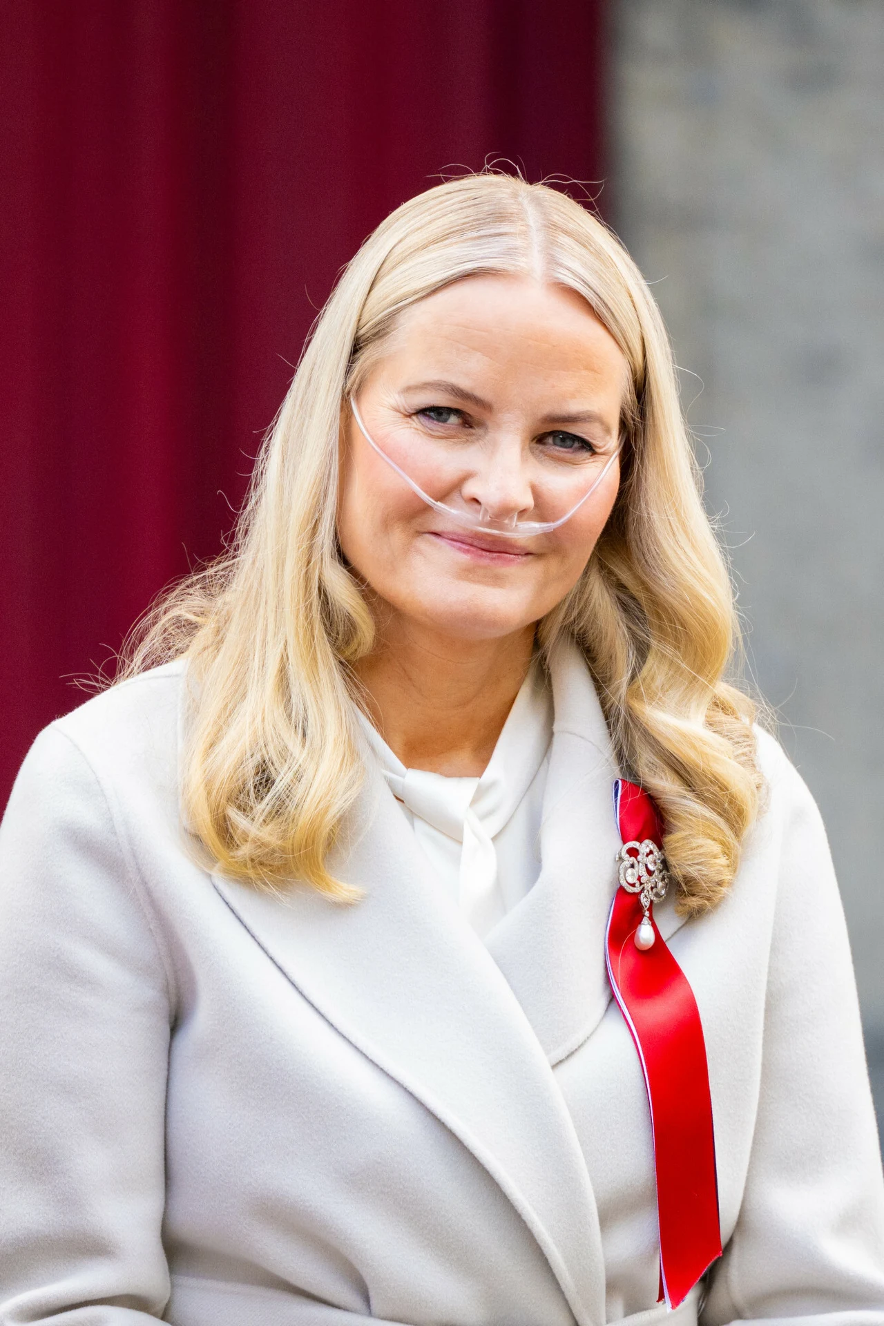 Uśmiechnięta kobieta z jasnymi, długimi włosami, ubrana w jasny płaszcz oraz białą koszulę, z tlenem do nosa i ozdobną czerwoną wstążką przypiętą do ubrania.