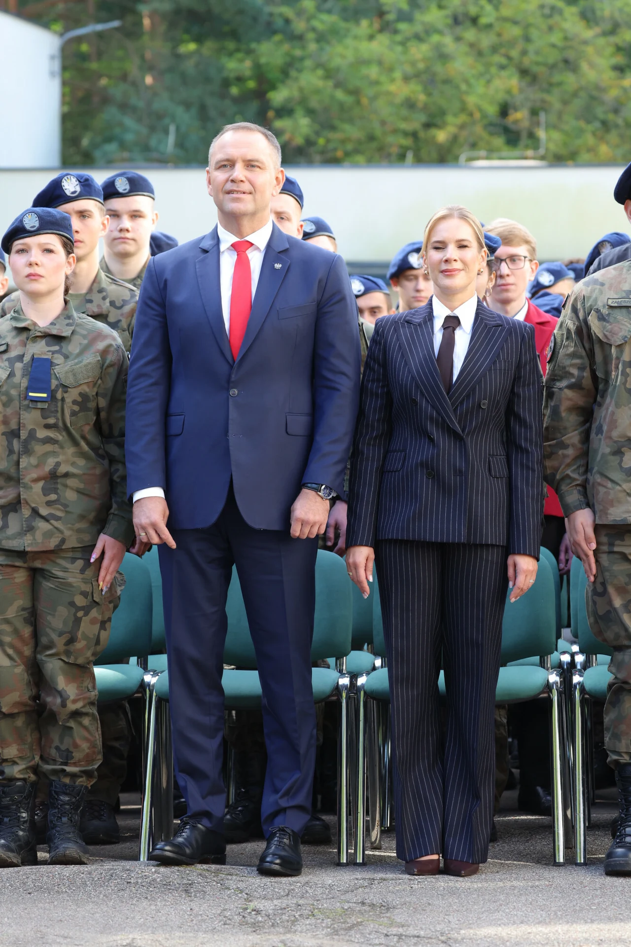 Grupa osób w mundurach wojskowych oraz dwie osoby w eleganckich garniturach stoją w pierwszym rzędzie podczas oficjalnego wydarzenia na świeżym powietrzu.