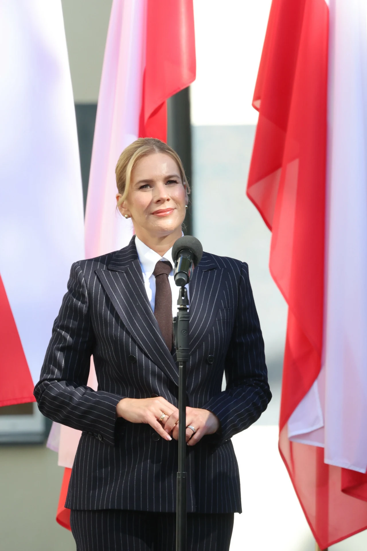 Kobieta w ciemnym, prążkowanym garniturze stoi przy mikrofonie na tle rozpostartych flag Polski.