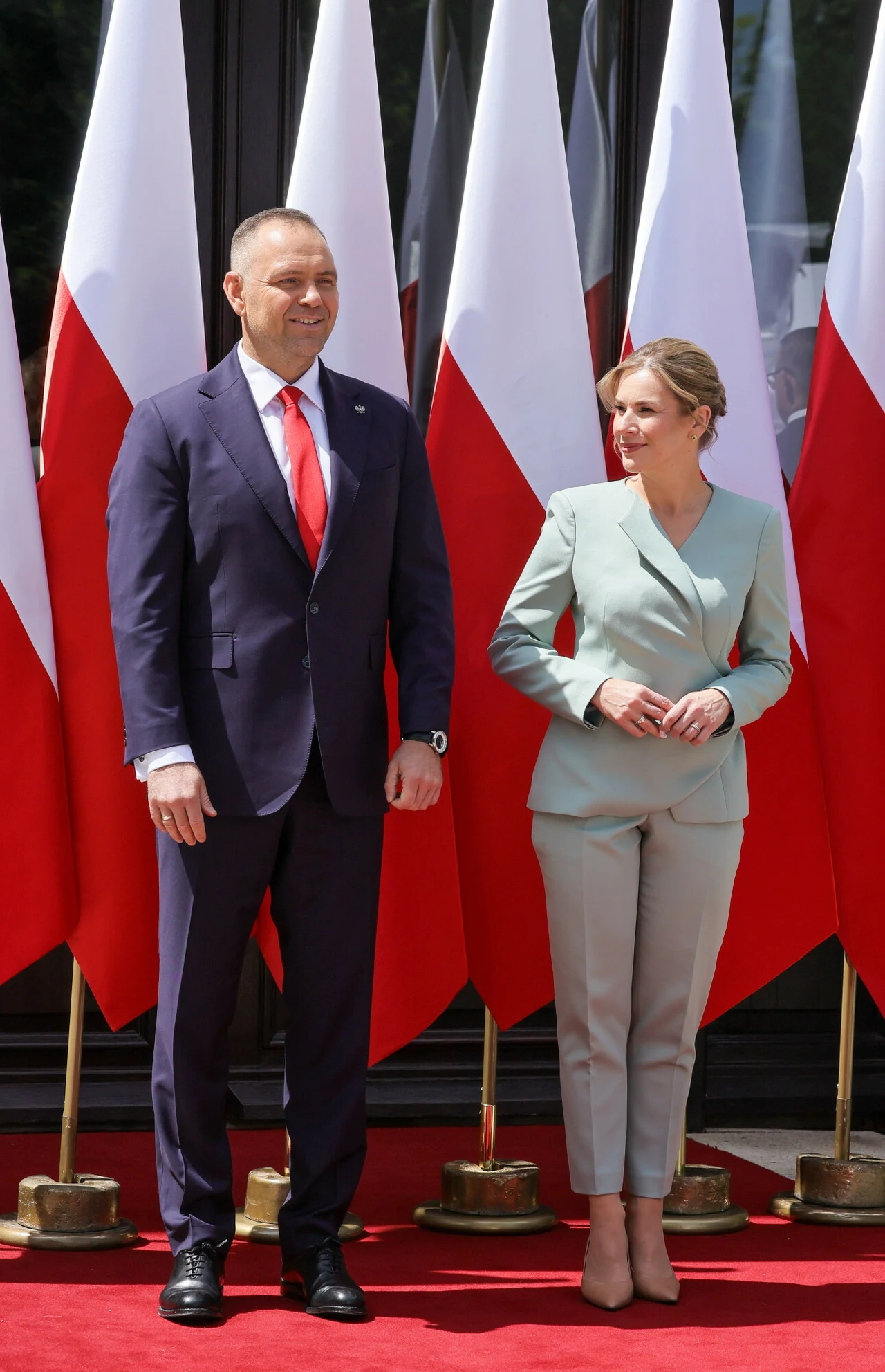 Dwoje elegancko ubranych osób stoi na czerwonym dywanie na tle ustawionych w rzędzie flag Polski.