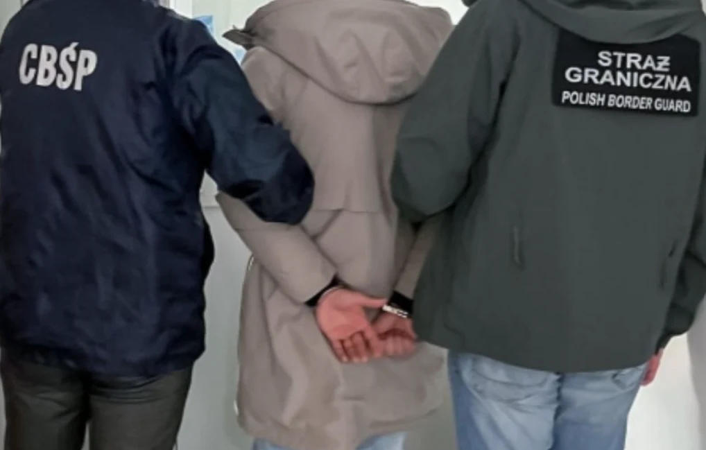 Osoba w jasnej kurtce z kajdankami na nadgarstkach stoi pomiędzy funkcjonariuszami Centralnego Biura Śledczego Policji i Straży Granicznej ubranymi w oznakowane kurtki służbowe.