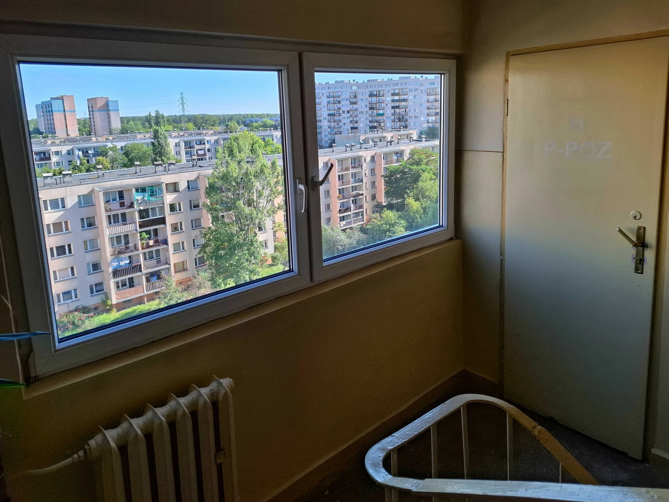 Widok na osiedle bloków mieszkalnych z okna klatki schodowej w wysokim budynku, z wyraźnymi oknami i drzwiami prowadzącymi na klatkę schodową.