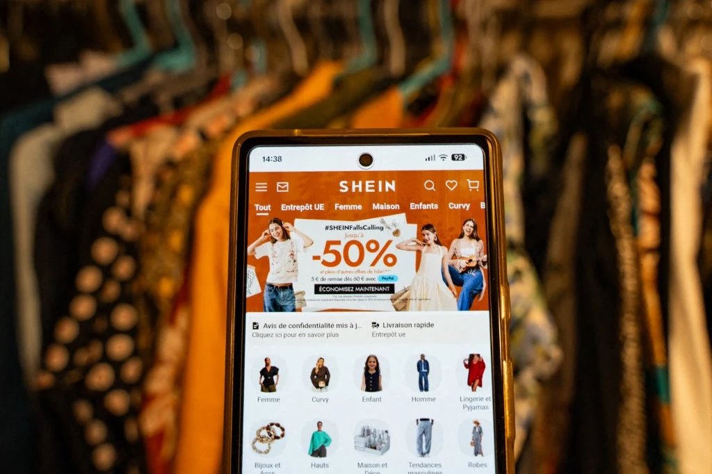 Od tego tygodnia Francja będzie nakładać karę na tanio wykonaną odzież Smartfon z aplikacją SHEIN pokazującą promocję -50% na tle garderoby z kolorowymi ubraniami na wieszakach.