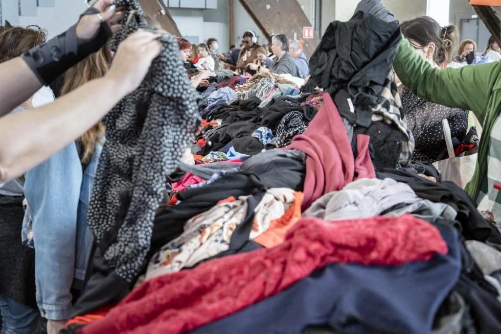 Ubrania z drugiej ręki nadal cieszą się powodzeniem Stół zasypany różnorodnymi ubraniami, wokół którego zgromadzone są osoby przeglądające odzież i wybierające interesujące je rzeczy.