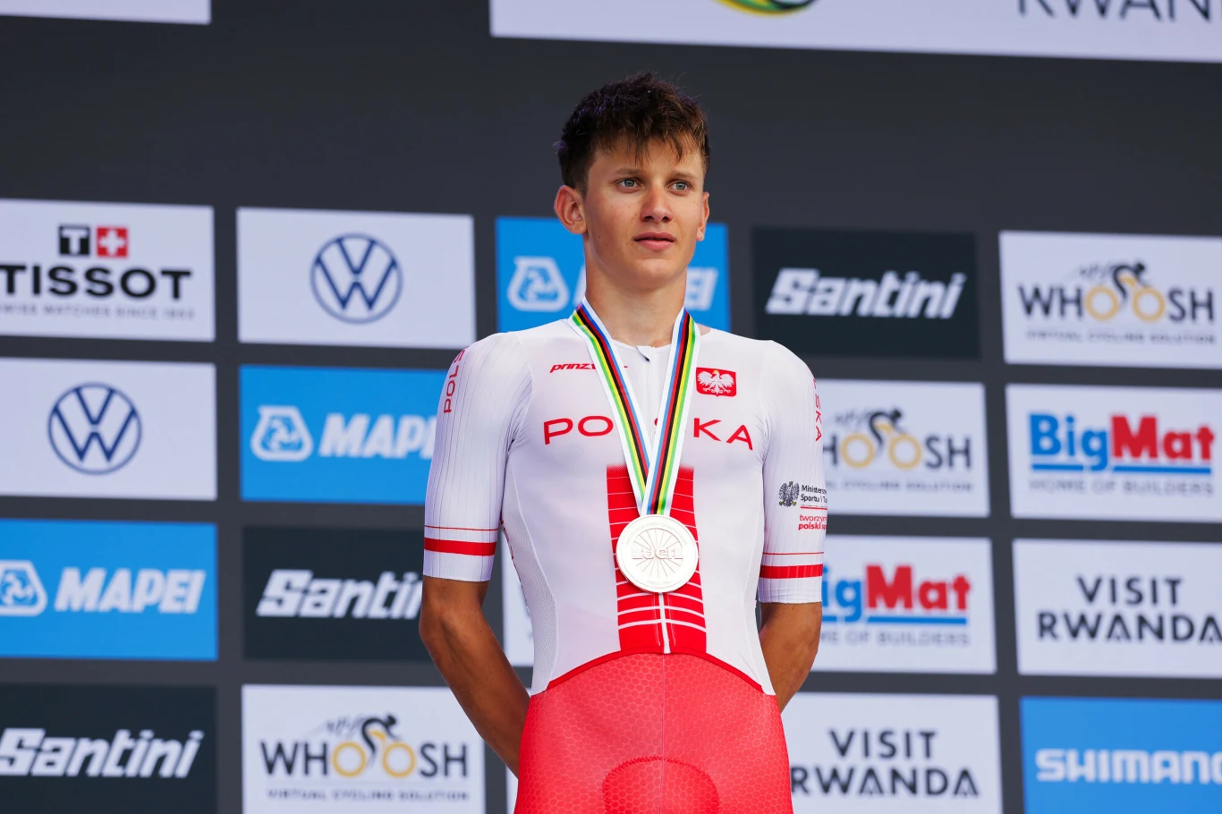 Polski kolarz w biało-czerwonym stroju z medalem na szyi stoi na podium na tle ściany z logotypami sponsorów wydarzenia sportowego.