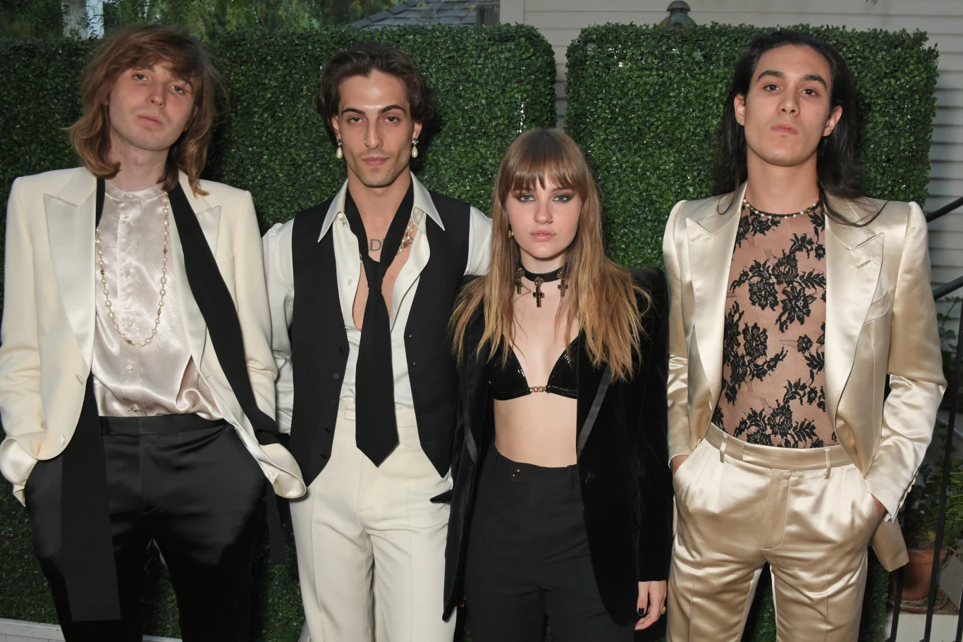Zespół Maneskin: Thomas Raggi, Damiano David, Victoria De Angelis i Ethan Torchio Zespół Maneskin w eleganckich garniturach i odważnych kreacjach na tle zielonego żywopłotu.