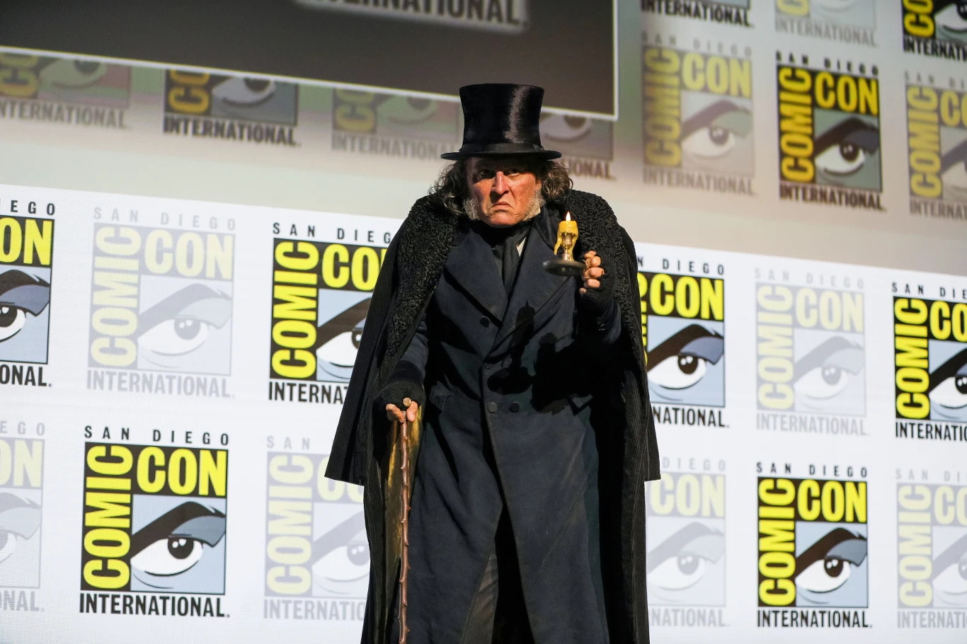 Mężczyzna w czarnym płaszczu i cylindrze trzyma laskę oraz statuetkę, stojąc na tle ściany z logotypami Comic Con International.