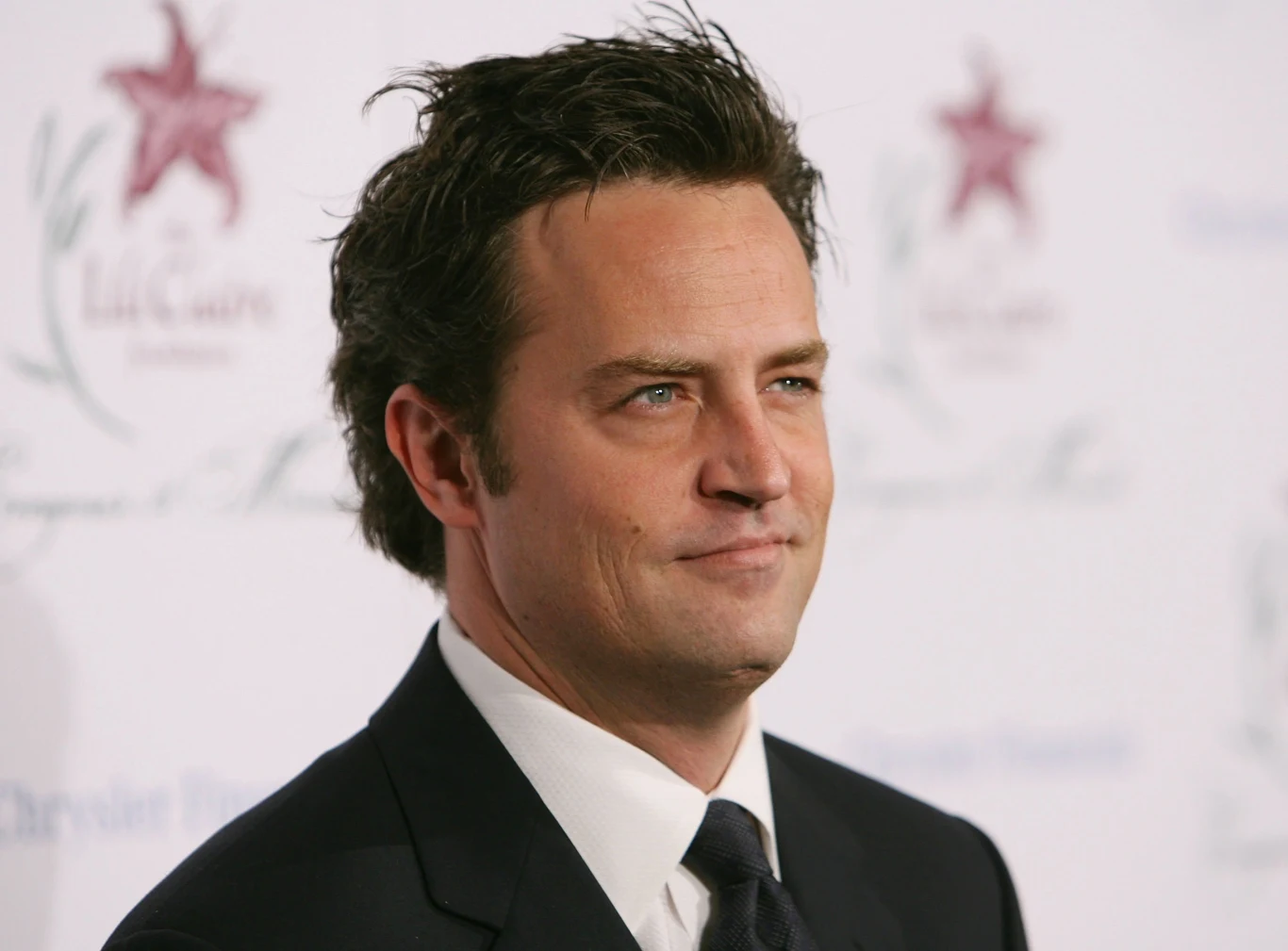Matthew Perry w 2006 roku Matthew Perry w 2006 roku. Mężczyzna w garniturze i białej koszuli na tle ściany z logo z czerwoną gwiazdą.