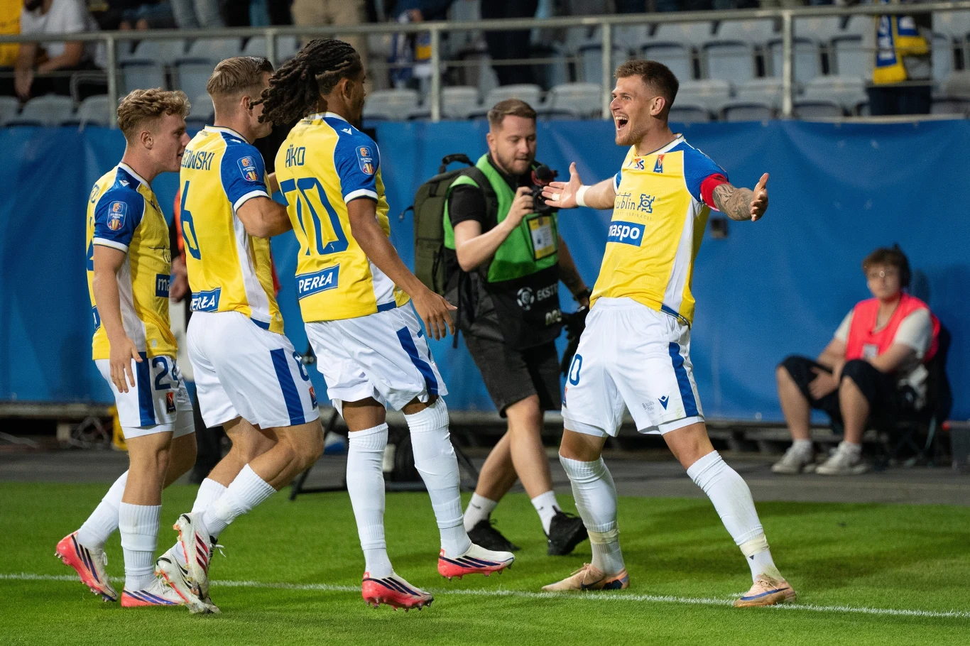 Karol Czubak otworzył wynik w meczu Motor Lublin - Legia Warszawa Czterech piłkarzy w żółto-niebieskich strojach celebrujących zdobycie gola na boisku, jeden z nich z rozłożonymi rękami biegnie w stronę reszty drużyny, a w tle widoczny jest fotoreporter i kibice na trybunach.
