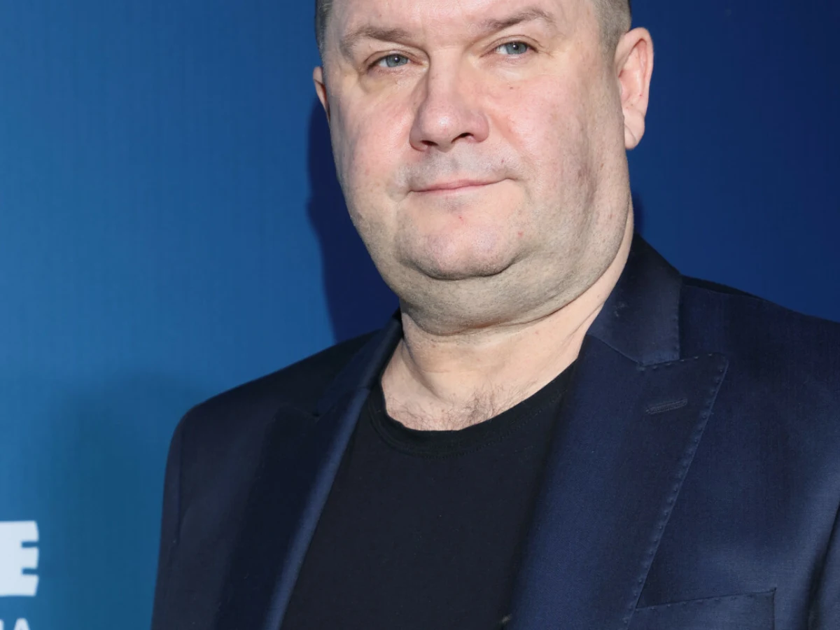 Mężczyzna w ciemnej marynarce i czarnej koszulce stoi na tle niebieskiej ścianki.