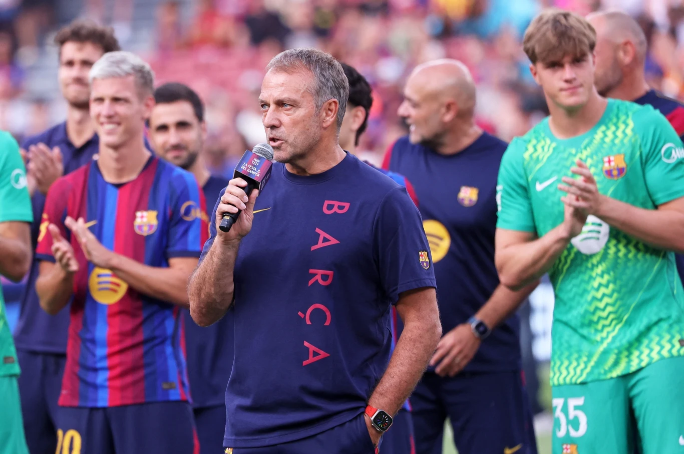 Hansi Flick Trener drużyny piłkarskiej Barcelona w granatowej koszulce z napisem BARÇA trzyma mikrofon, otoczony przez zawodników w strojach meczowych, którzy stoją i klaszczą.